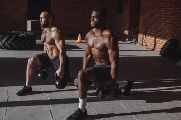 Two multiracial male athletes making lunges with weight in indoor workout