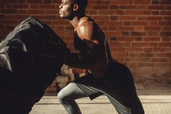 Atlético afroamericano hombre haciendo ejercicio con neumático y mirando hacia otro lado en el gimnasio —  Fotos de Stock