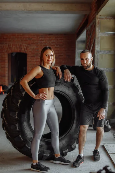 Young trained woman working out with truck tire with personal trainer assistance