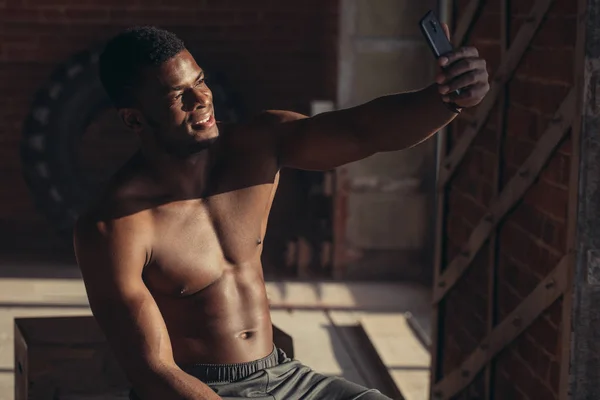 Vista lateral del atleta afroamericano sin camisa descansando y usando un teléfono inteligente en el neumático en el gimnasio —  Fotos de Stock