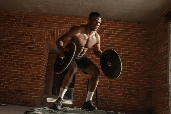 Hombre africano sin camisa con placas de barra salta en el aire en el neumático del camión en el gimnasio —  Fotos de Stock