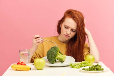 Vegetable diet. Sad dull woman holding broccoli on fork while making grimace clipart