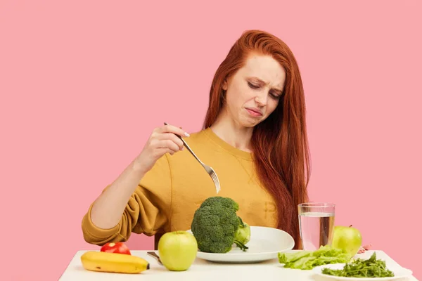 Zeleninová strava. Smutná tupá žena drží brokolici na vidličce, zatímco se šklebí — Stock fotografie