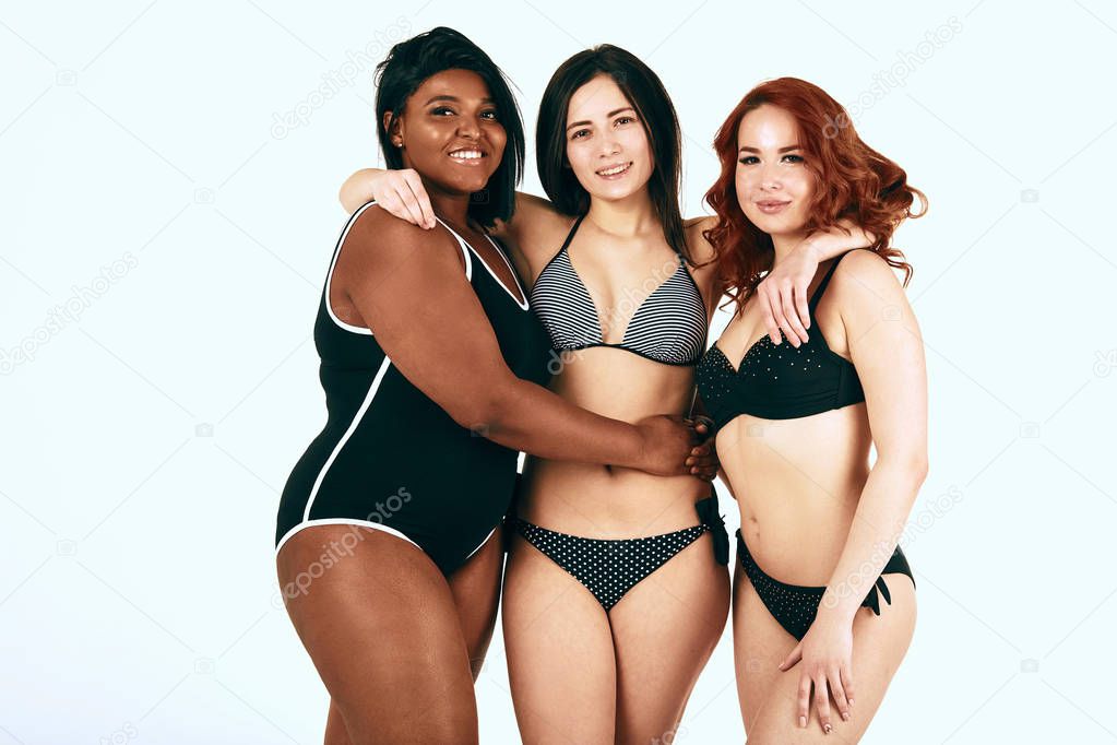 Multiracial group of three happy diverse women in swimsuits hugging on white