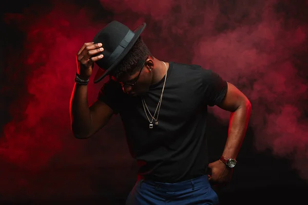 Ballerino africano con cappello che balla in t-shirt nera in club fumoso rosso scuro — Foto Stock