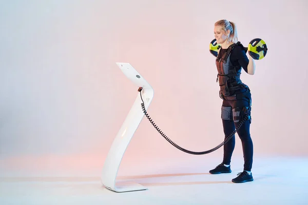 Nice blonde girl wearing EMS costume working out with barbell indoors. — Stock Photo, Image