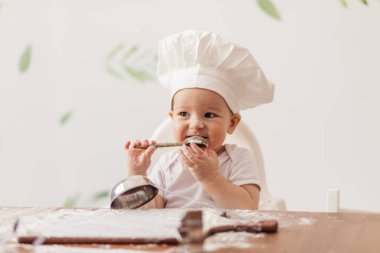 Mutfak masasında hamurla oynayan aşçı şapkası takan bebek portresi.