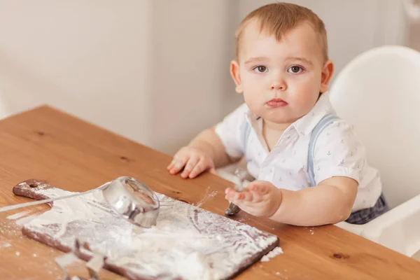 Menino fogão infantil ajuda a fazer bolos sentados à mesa com farinha espalhada. — Fotografia de Stock