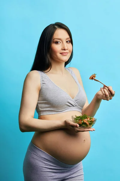 Femeie gravidă subțire cu burtă mare mănâncă salată de legume izolată peste peretele albastru — Fotografie, imagine de stoc