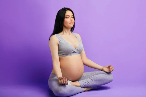 Femeia însărcinată mediază și ține ochii închiși în timp ce stă în poziția de lotus — Fotografie, imagine de stoc