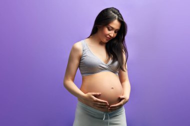 Mor duvarın üstünde çıplak karnına poz veren hamile bir kadın.