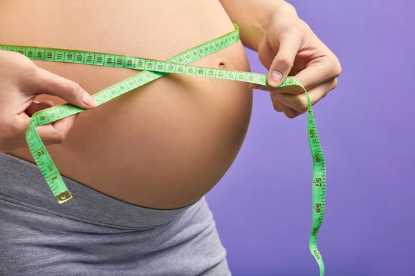 Έγκυος γυναίκα σε croptop μετρά γυμνή κοιλιά της με ταινία μέτρησης στο μωβ — Φωτογραφία Αρχείου