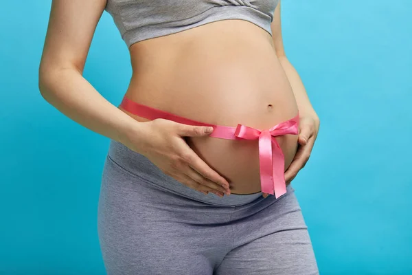 Beautiful pregnant woman tying up her tummy with pink ribbon — Stock Photo, Image