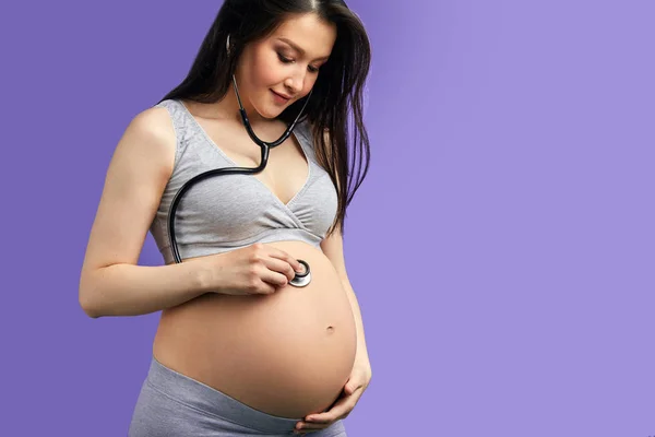Mujer embarazada sonriente escuchando su vientre usando un estetoscopio — Foto de Stock