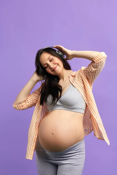 Mujer embarazada sonriente en auriculares bailando en estudio sobre fondo morado . — Foto de Stock