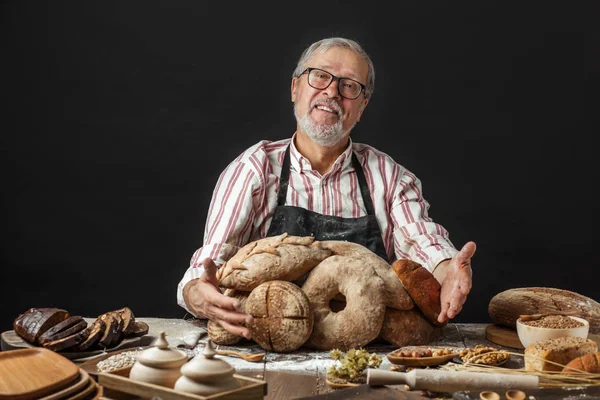 Feliz velho padeiro olhando para a câmera e sorrindo enquanto abraça pães — Fotografia de Stock