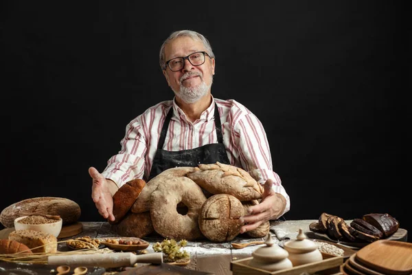 Счастливый старый пекарь смотрит в камеру и улыбается, обнимая буханки хлеба — стоковое фото