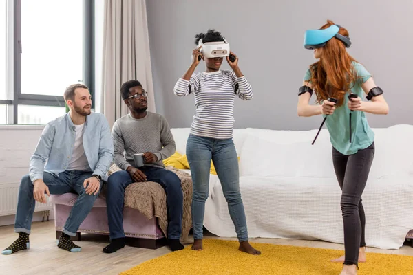 Two multiracial young women dancing wearing virtual reality glasses while their boyfriends sitting on couch and smiling