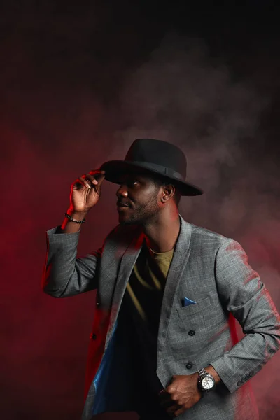 Afrikanischer Geschäftsmann in grauem Anzug und schwarzem Hut im Studio auf dunklem Hintergrund — Stockfoto