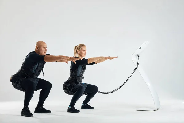 A couple wearing suits for electro muscle stimulation EMS performing squats — Stock Photo, Image