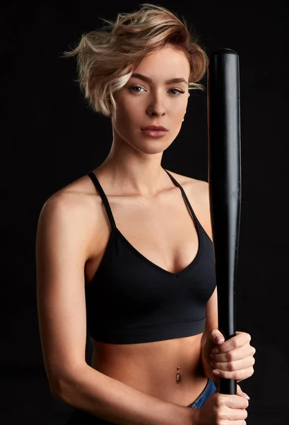 Sexy slank fit meisje in zwarte top houdt honkbalknuppel. Geïsoleerd over zwarte achtergrond. — Stockfoto