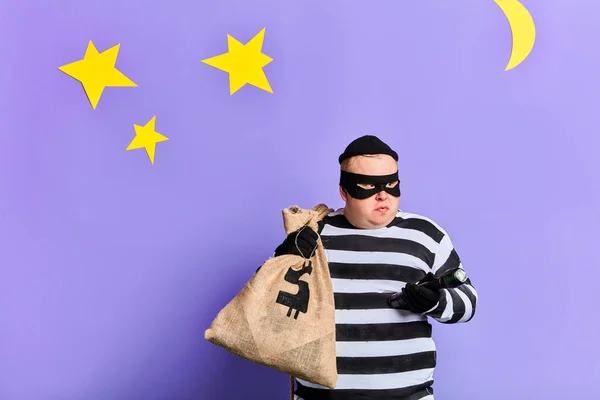 Büyük bir çanta taşıyan ciddi şişman hırsız, çizgili giysiler giymiş, maske — Stok fotoğraf