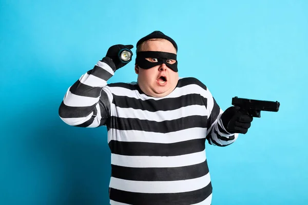Desconcertado regordete ladrón sosteniendo un arma en un fondo azul —  Fotos de Stock