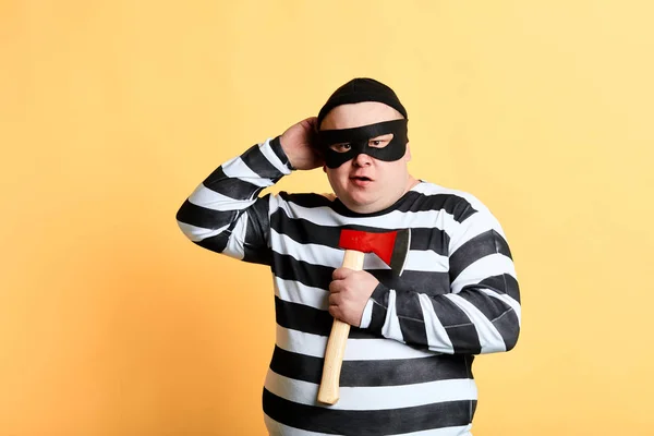 Pummeliger Verbrecher mit Maske, der sich den Kopf kratzt und die Axt in der Hand hält. Studioaufnahme. — Stockbild Dicker Verbrecher mit Maske, der sich am Kopf kratzt, Axt hält — Stockfoto