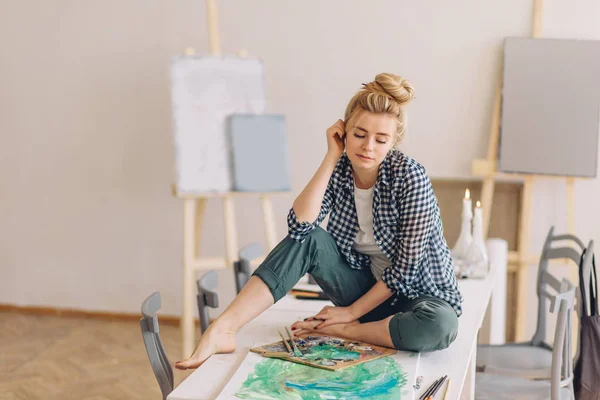 Ładna kobieta siedzi na stole w pobliżu palety w studiu sztuki — Zdjęcie stockowe