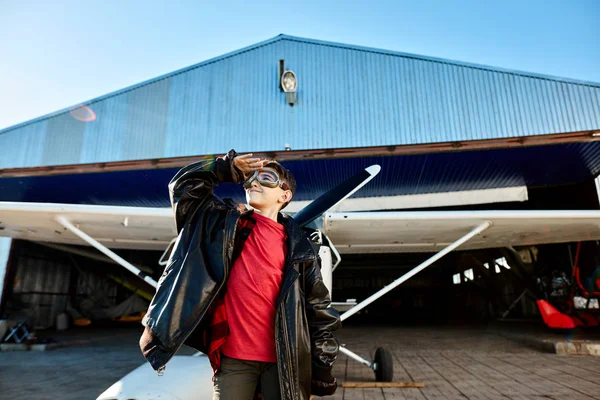 Niño se para cerca del hangar, sombrea los ojos con la mano, mira al cielo con sonrisa — Foto de Stock