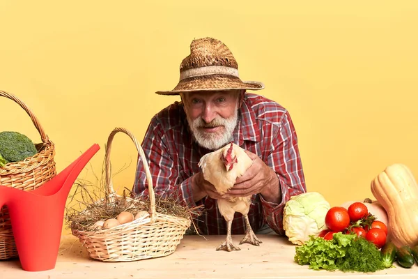 Agricultor com barba cinza captura galinha, de pé atrás da mesa de madeira — Fotografia de Stock