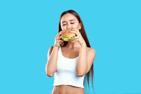Sexy atlética mujer disfrutando de comer hamburguesa sobre fondo azul —  Fotos de Stock