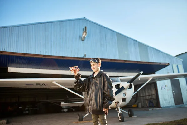 Lindo niño de pie jugando con el modelo de avión, lleva chaqueta de piloto grande — Foto de Stock