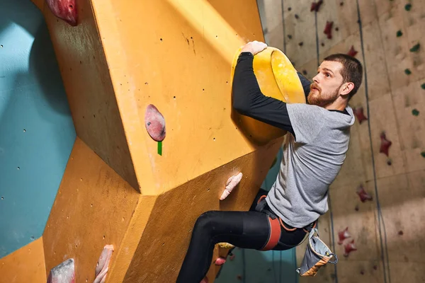 Handsome boulderer hanging on large hold at colorful climbing wall — Stock Photo, Image