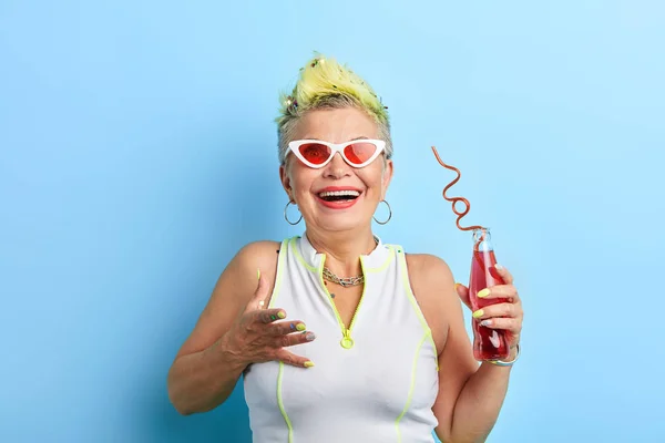 Senhora idosa com cabelo amarelo curto em roupa engraçada beber coquetel vermelho — Fotografia de Stock