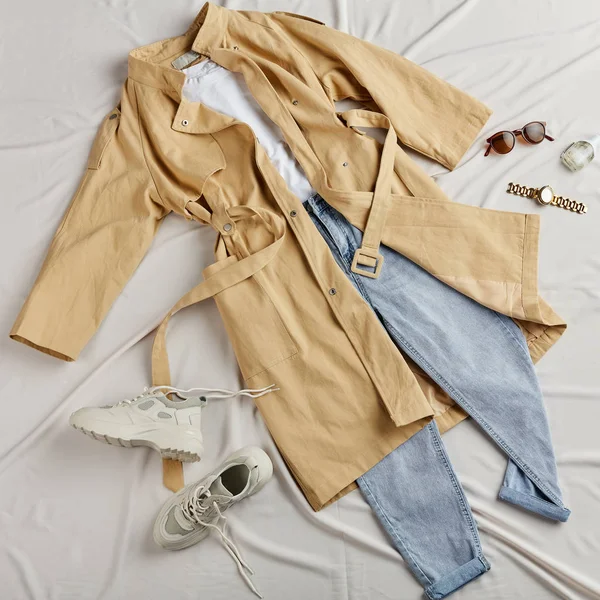 Moderne beige frakk, jeans og hvite trenere som ligger på det hvite arket – stockfoto