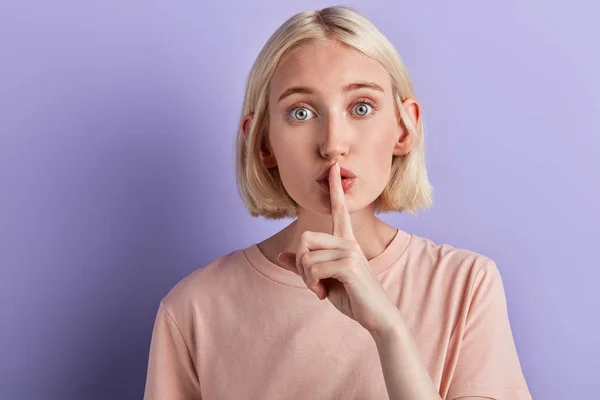 Blonde girl showing shhh sign with finger to lips over violet background — Stock Photo, Image