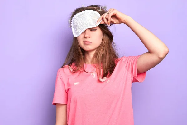 Exhausted young woman raises slightly her eye mask, trying to open sleepy eyes