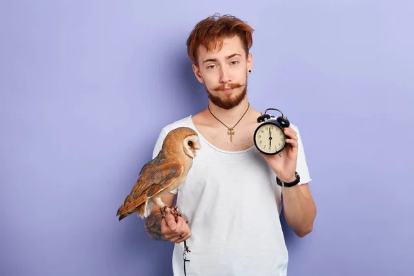 Sleepy unhappy man with an owl on his arm holds alarm clock