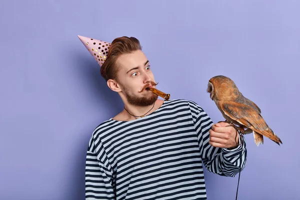 Homem engraçado positivo no chapéu e com chifre de festa em sua boca olhando para o pássaro — Fotografia de Stock