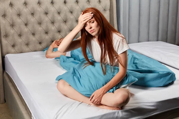 Pobre chica pelirroja sentada en la cama tocándose la frente — Foto de Stock