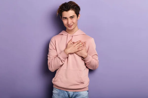 friendly positive asian guy in pink hoodie touching his heart with palms