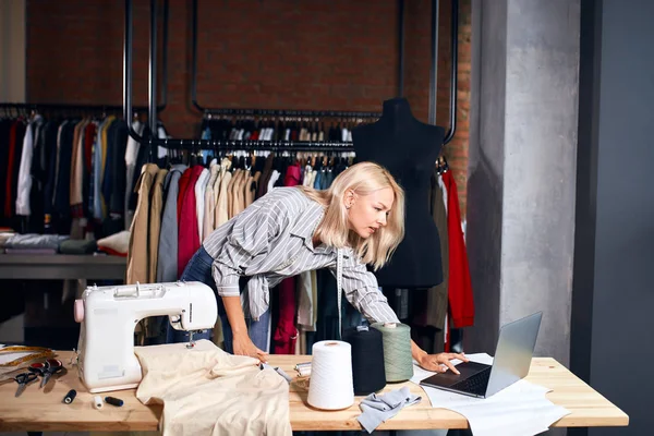 Молодая блондинка портной с помощью ноутбука во время шитья одежды — стоковое фото