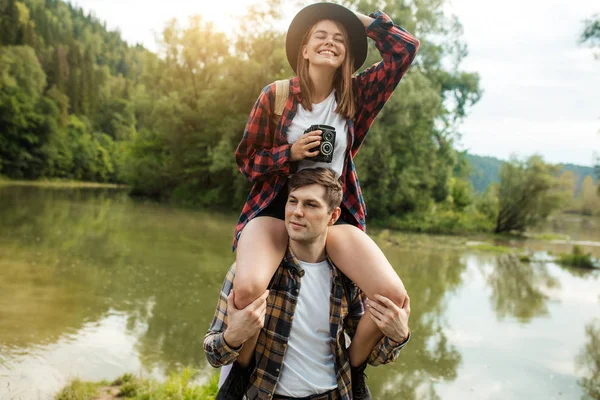Fair-haired girl in checked shirt and hat laughing, enjoying sitting on guys shoulder — Stock Photo, Image