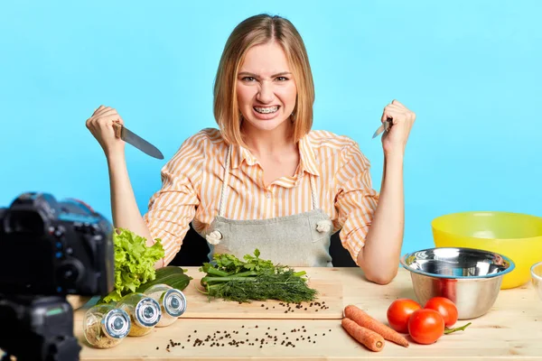 Blonde female cook holding two knives in hands, has mad face expression