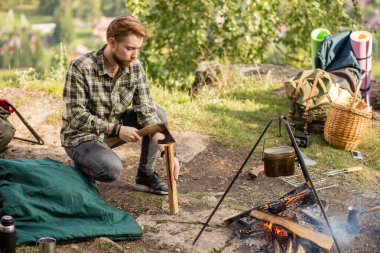 Genç sakallı çekici turist, yürüyüşçü şenlik ateşine odun hazırlıyor.