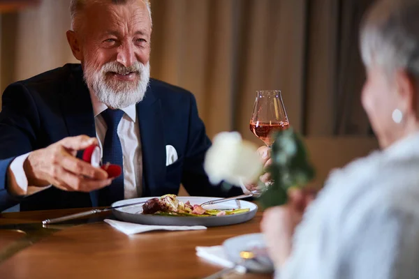 Όψη πάνω από τον ώμο του ώριμου άνδρα με σμόκιν πρόταση γάμου σε γυναίκα κρατώντας λευκό τριαντάφυλλο στο εστιατόριο — Φωτογραφία Αρχείου