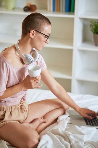 Biała dziewczyna ze słuchawkami oglądającymi w laptopie na łóżku z filiżanką herbaty — Zdjęcie stockowe