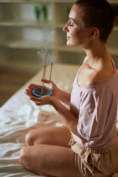 Beautiful caucasian girl meditating while sitting on bed with burning aroma incense sticks — Stock Photo, Image