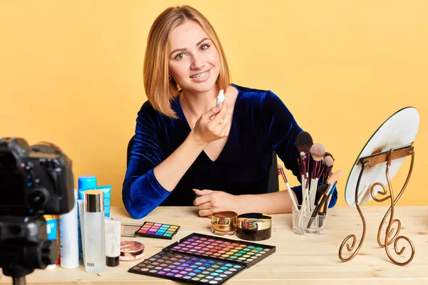 Cute blonde vlogger sits at table surrounded with cosmetics, smiles broadly — Stock Photo, Image